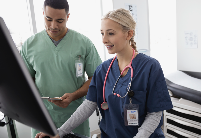 A male nurse in green scrubs looking at a clipboard and a female nurse in blue scrubs using a computer