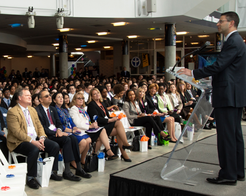 A man in a suit standing at a lectern in front of a full auditorium