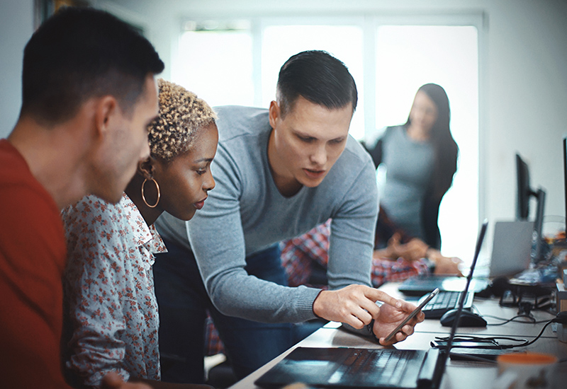 Three young people at a table with laptops looking at a smartphone together
